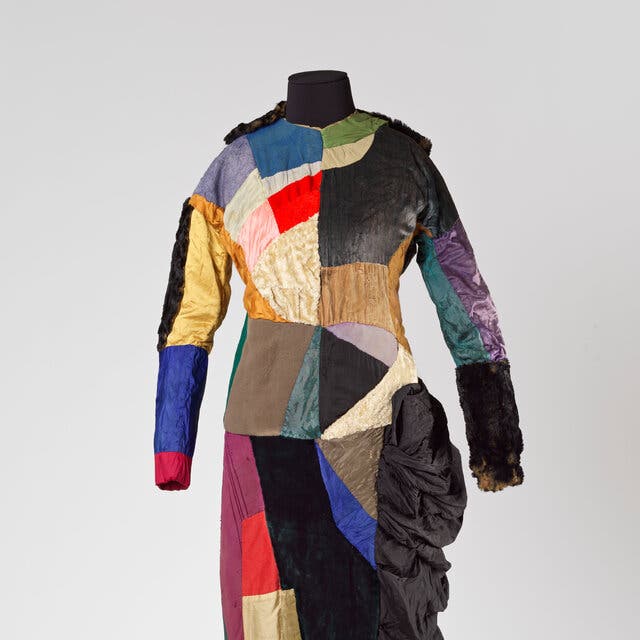 A patchwork dress sewn from various fabric pieces, in red, blue, yellow, green and black hues, installed on a stand.