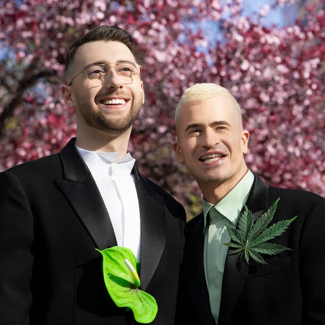 A couple stand together outdoors and smile. Billy Jacobson wears a black suit, a white dress shirt and a handmade anthurium brooch. Evan Ross Katz wears a black suit, a green dress shirt and a handmade marijuana leaf brooch. 