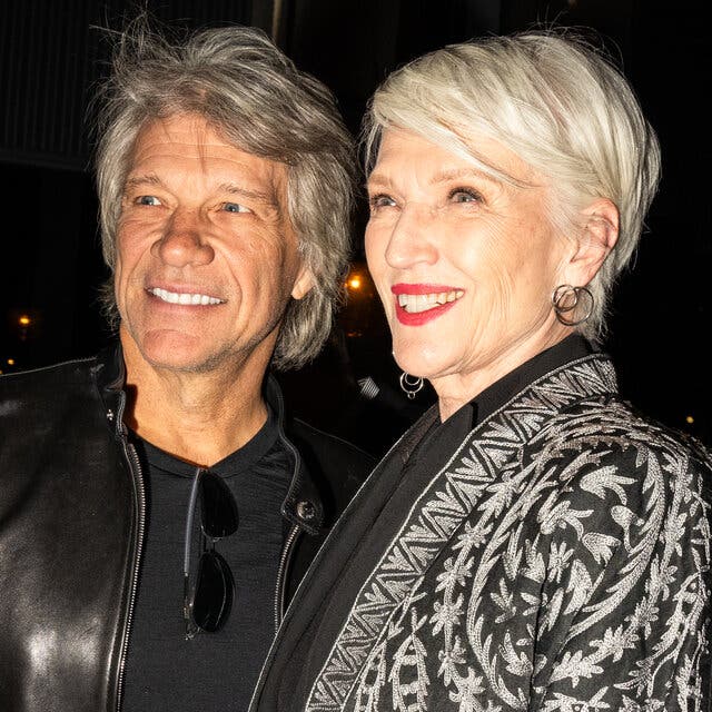 Jon Bon Jovi, wearing a leather jacket over a black T-shirt, and Maye Musk, wearing a patterned black-and-white jacket, stand together at a party, smiling.