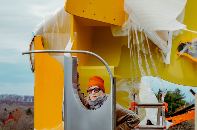 Arlene Shechet with the 20-foot-tall “As April,” one of six massive welded sculptures from “Girl Group,” her exhibition being installed at Storm King Art Center. The 2024 work is in two vivid shades of yellow. 
