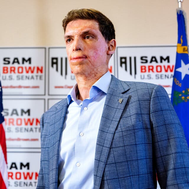 Sam Brown, in a blue shirt and blue blazer, stands in front of a wall decorated with his campaign signs.