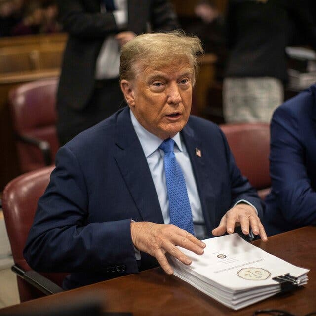 Donald Trump speaks at a defendant’s table with his hands atop a stack of paper. His lawyer Todd Blanche sits to his left.
