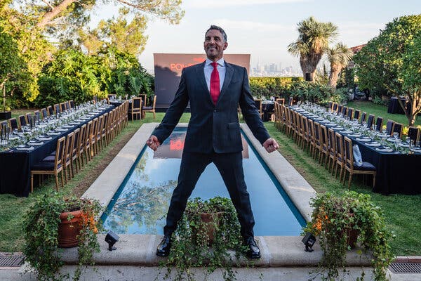 Mauricio Umansky, in a black suit and a red tie, stands on the edge of an infinity pool with a banquet set behind him in a tree-lined yard. He is grinning and posing in a wide stance with his fists by his sides.
