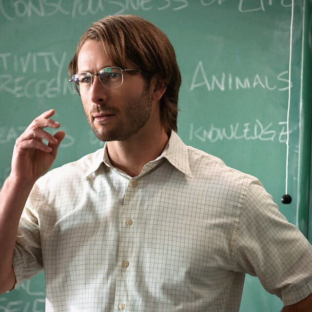 In a movie scene, a nerdy looking man in glasses and shirt sleeves stands in front of a green chalkboard with words like “subjectivity” and “knowledge” written on it.