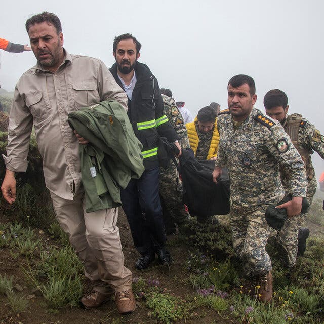 Men in military and civilian dress carrying a body in a black bag on a foggy mountain.