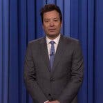 Jimmy Fallon, in a dark gray suit and purple tie, stands onstage in front of a blue curtain.