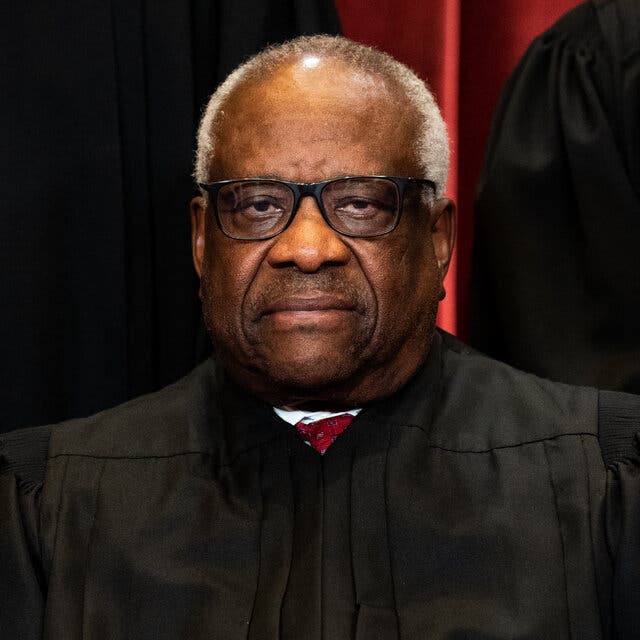 Justice Clarence Thomas, wearing a black robe, glasses and a red tie, sitting for a Supreme Court portrait.