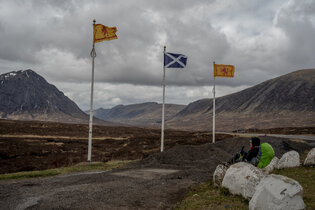 A camper in an mountainous area of Scotland, sitting next to flagpoles flying the country's blue-and-white Saltire flag and its red-and-yellow Rampant Lion flag.