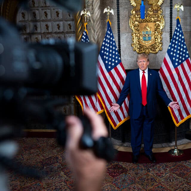 Donald Trump speaks to a television camera and stands between American flags. 