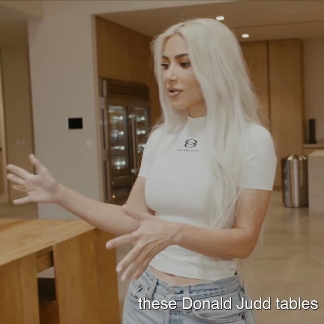 A woman with white hair in a white shirt and jeans stands next to a table.