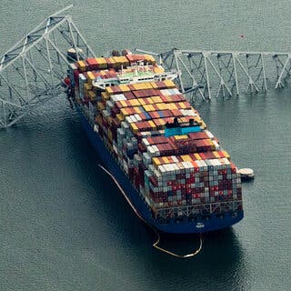 A large cargo ship is seen from above after it crashed into a bridge. The bridge is partly collapsed into the water.