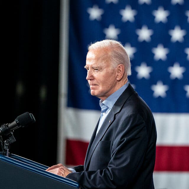 President Biden stands at a lectern in profile, speaking into two microphones. A large American flag is in the background.