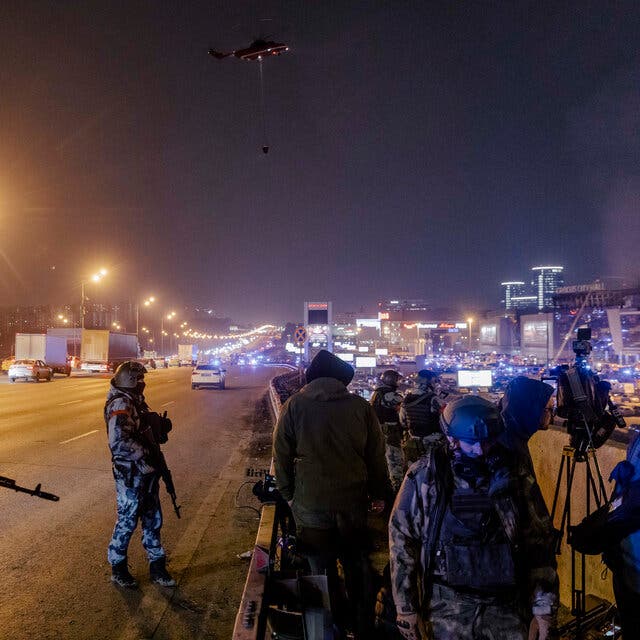 Two masked, heavily armed security personnel standing on a large road at night.