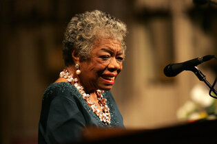 A smiling elderly Black woman in a turquoise dress and a floral necklace stands in front of a microphone.