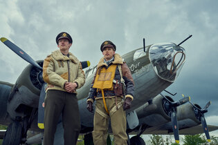 Two men in brown leather World War II aviator uniforms standing in front of a big silver plane.