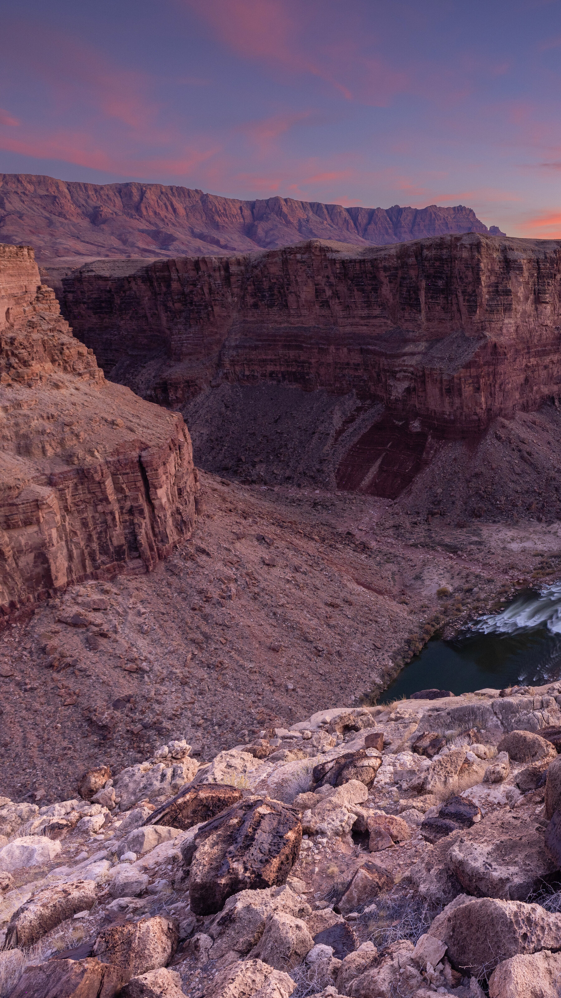 A wide river runs through the base of an immense canyon, which steeps cliffs leaving down to the water far below. The sky is a gradient of blues and pinks, and the light colors the canyon walls, giving the entire picture a rich and warm tone.