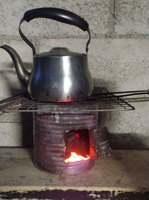 A teapot sitting on top of a metal grate, placed on a stove made of a large can with a cutout for adding wood.