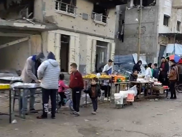A few vendors stand behind tables displaying food and other supplies for sale on the side of the road. Several adults and children survey the items. Behind the tables is a heavily damaged building and a building riddled with bullet holes.
