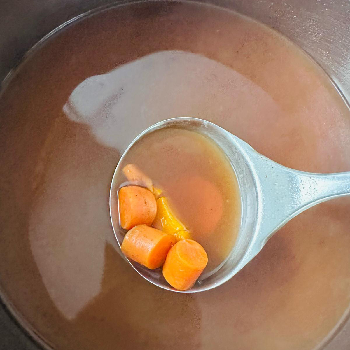 A pot of thin broth with pieces of carrot visible in a spoon.