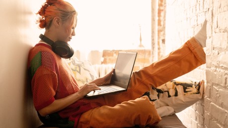Uma jovem com cabelo laranja senta-se confortavelmente perto de uma janela com fones de ouvido over-the-ear em torno de seu pescoço olhando para seu laptop Windows 11.